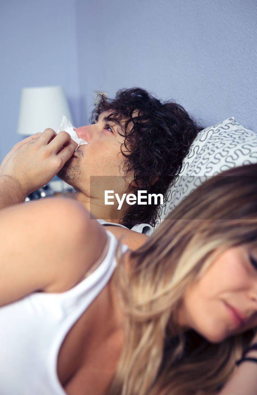 Young man blowing nose with girlfriend sleeping on bed at home