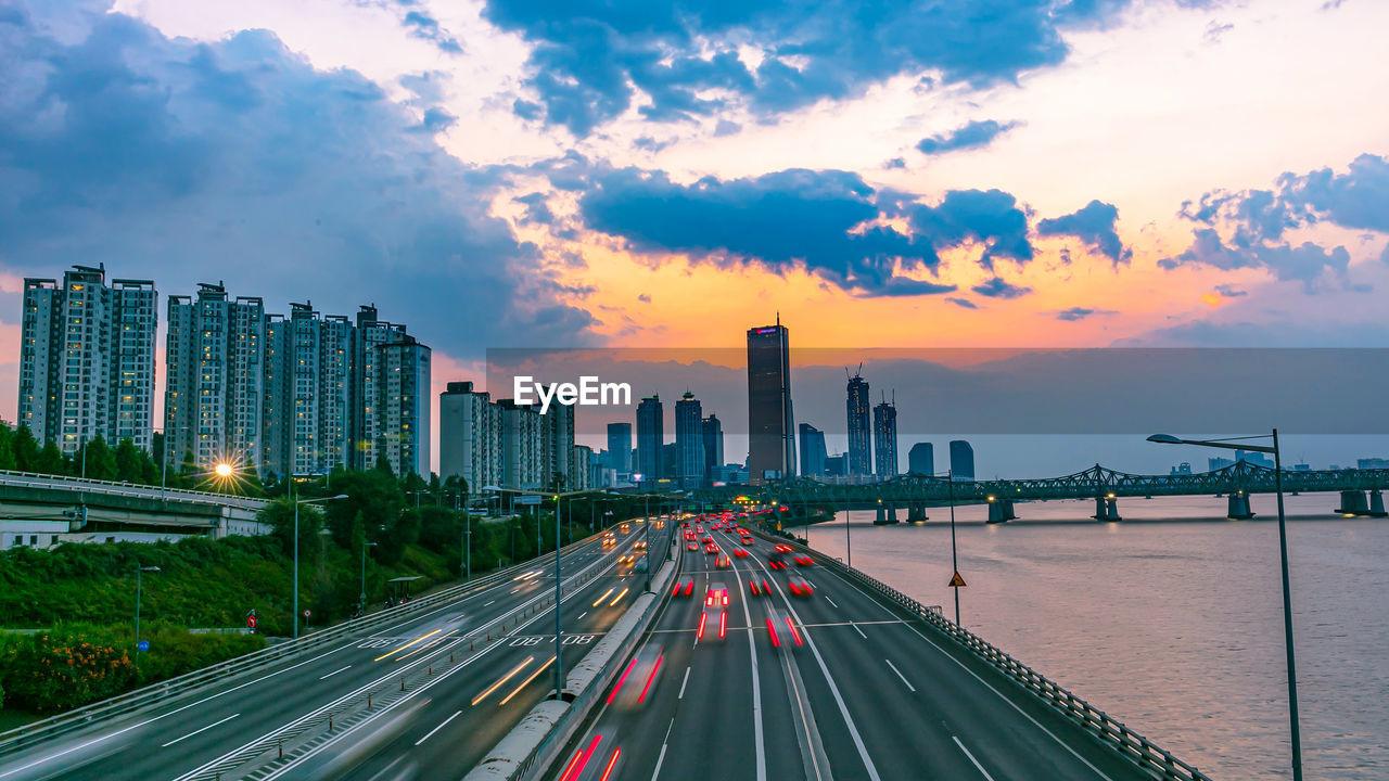 Light trails on road by river against sky during sunset