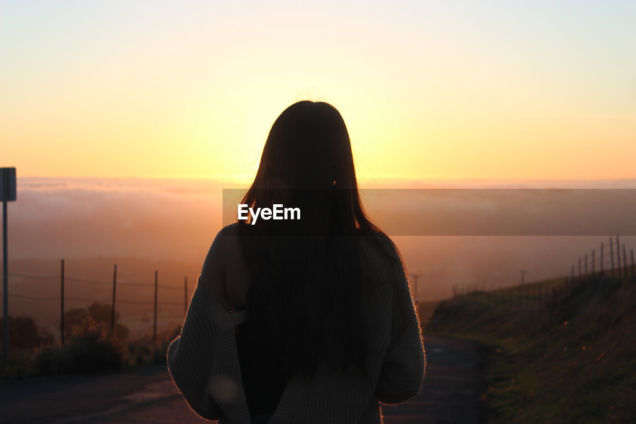 Rear view of woman wearing sweater standing on road against cloudscape during sunrise