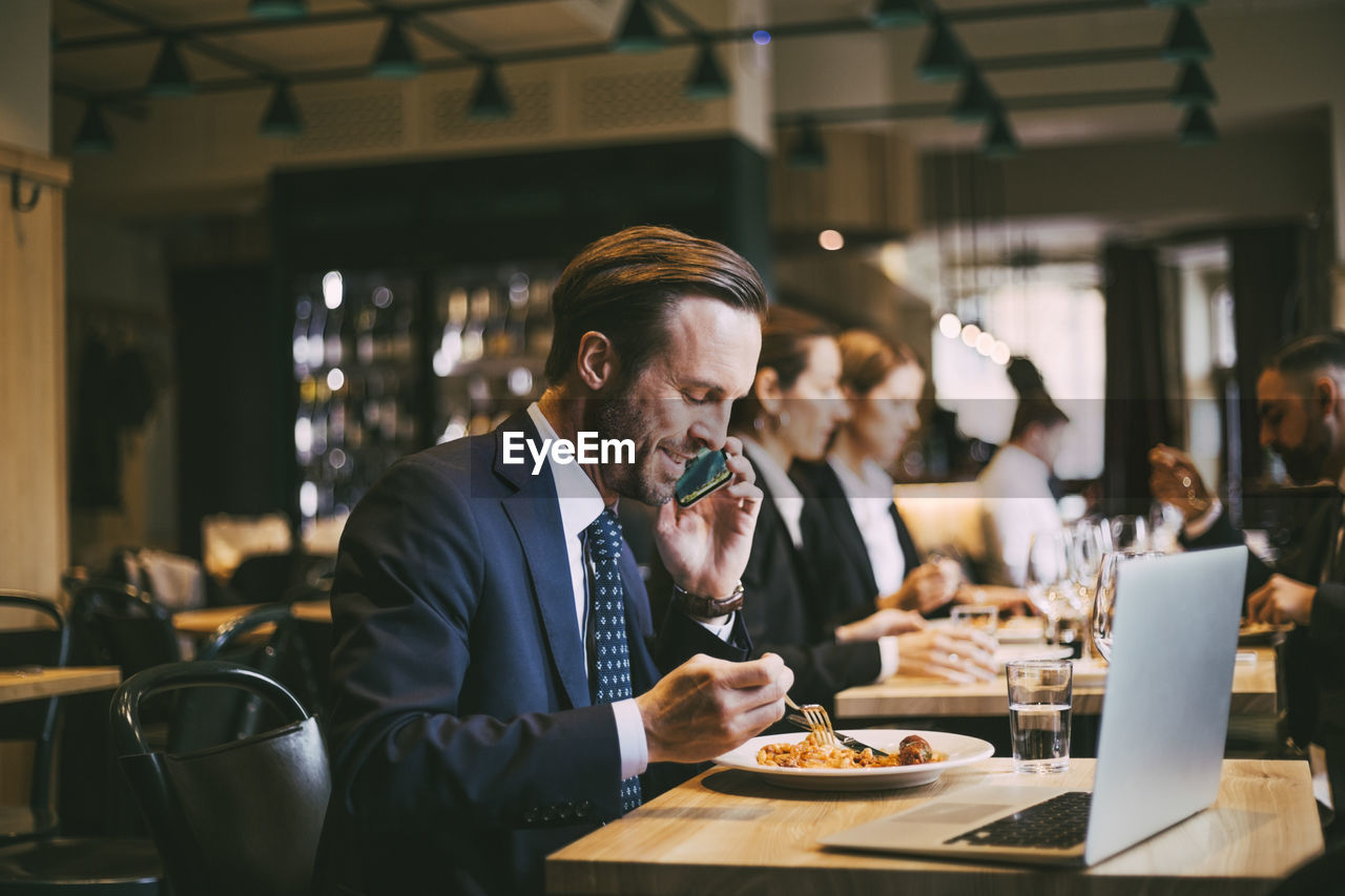 Smiling male business person talking on phone while eating food in restaurant