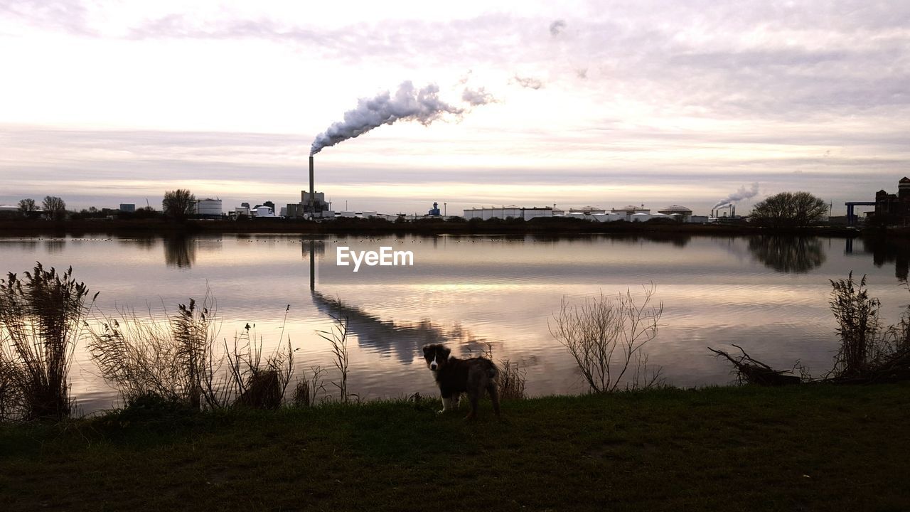 Smoke emitting from stack in front of river against sky during sunset