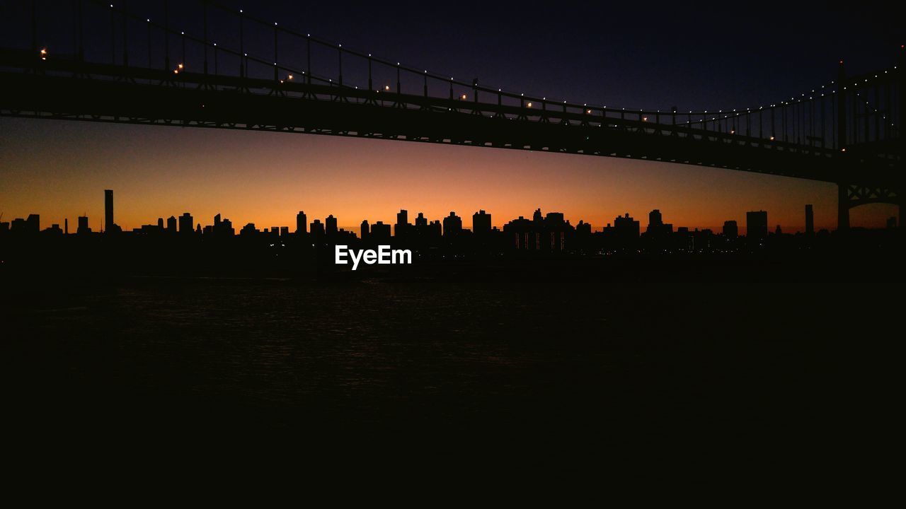 Illuminated silhouette bridge over river against sky at dusk