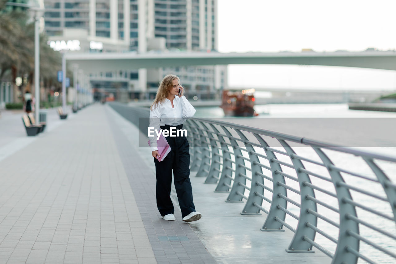 A blonde businesswoman holds a folder and talks on her smartphone in the city