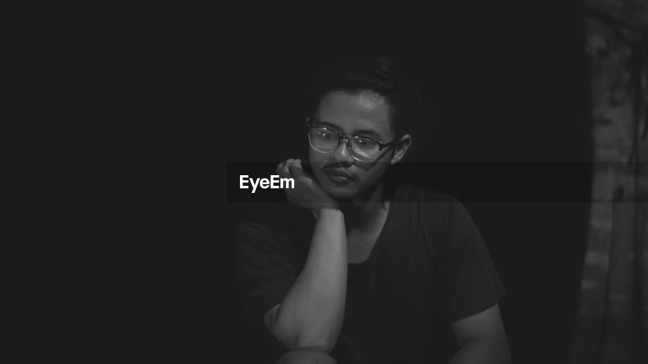 Thoughtful young man wearing eyeglasses sitting against black background