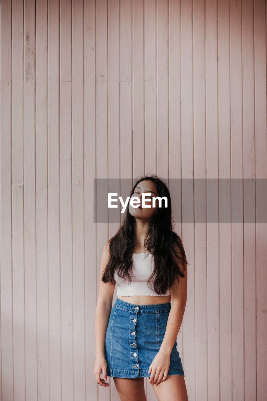 Calm dreamy female teenager with long dark hair wearing casual crop top and denim skirt standing with eyes closed against plank wall