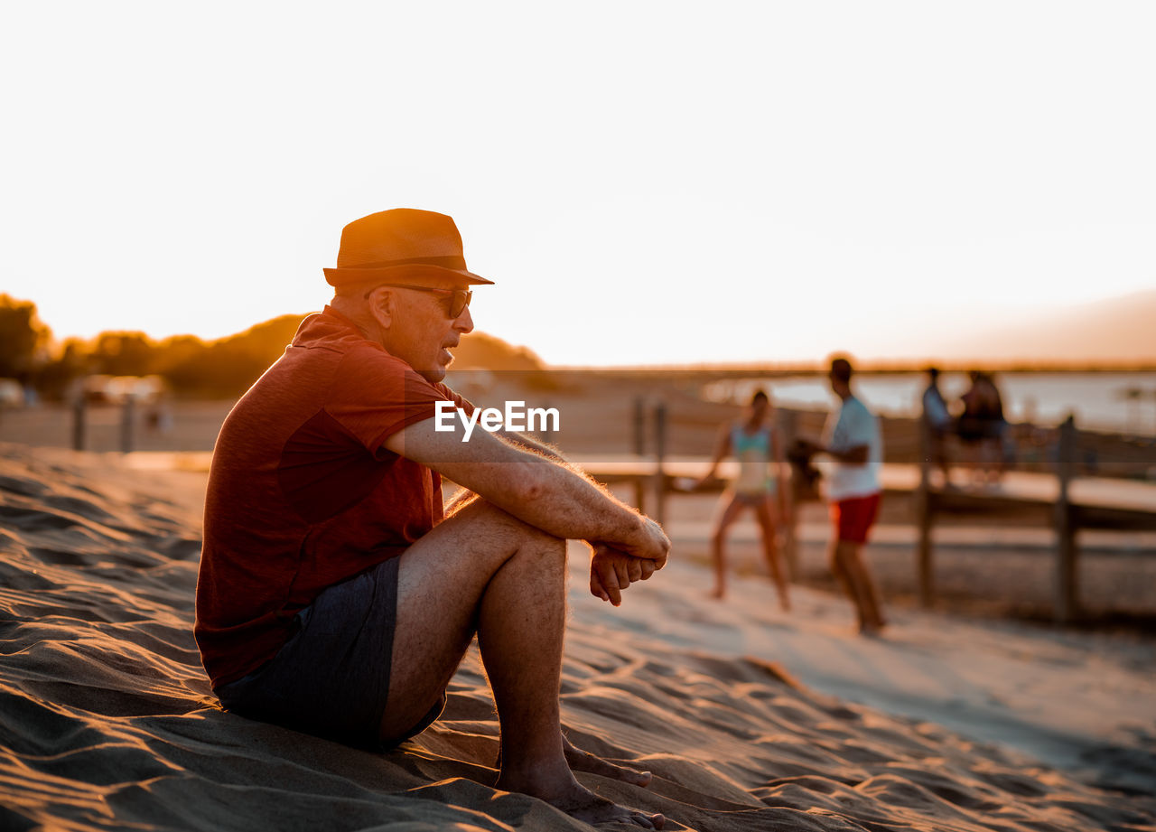 Side view full length contemplative adult male in summer wear resting on warm sandy beach at sunset