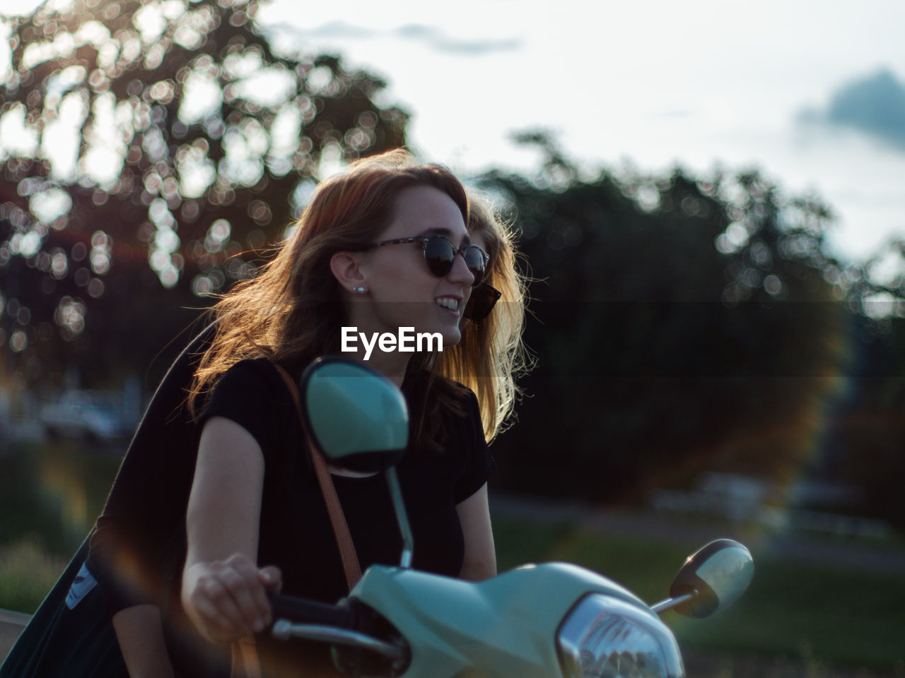 Young woman wearing sunglasses sitting on motor scooter against trees