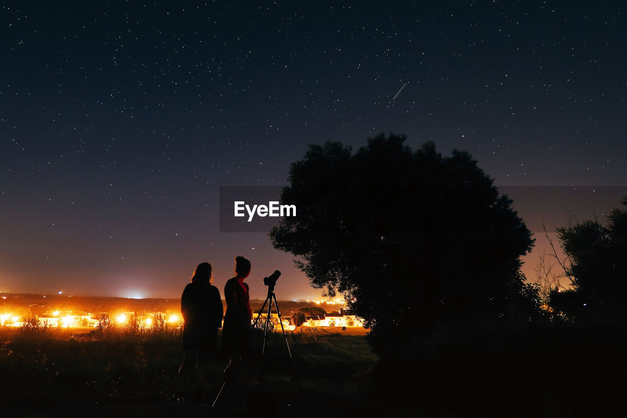 Silhouette female friends standing with telescope on field against star field