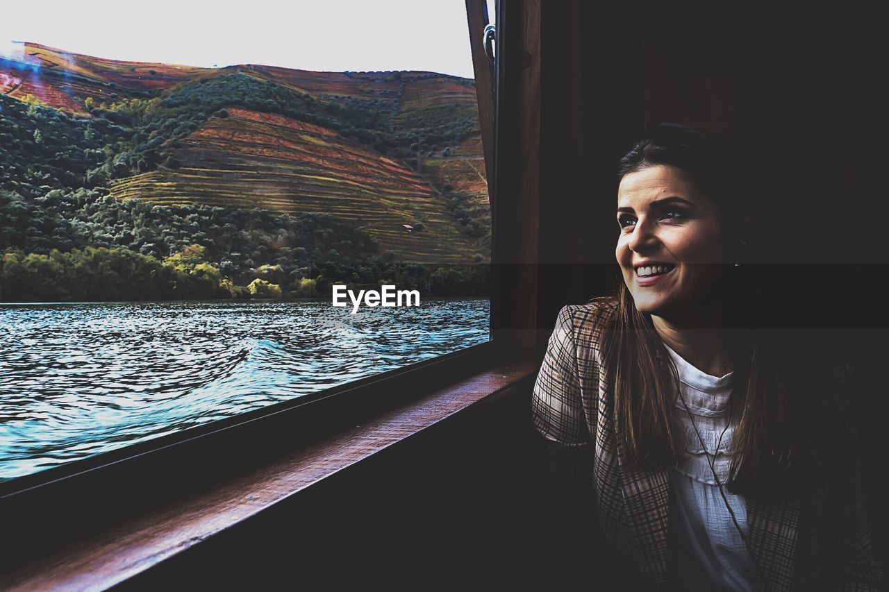 Smiling young woman looking through window at lake