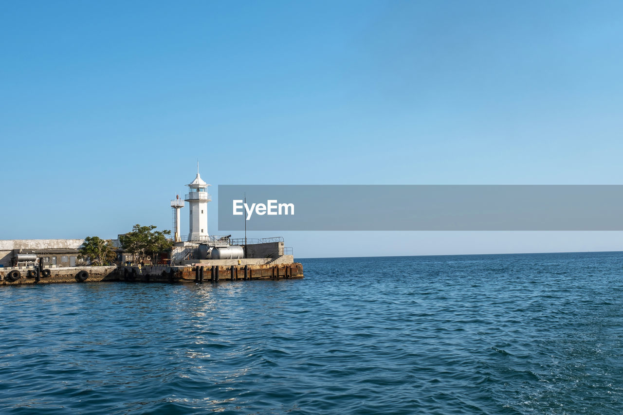 Lighthouse by sea against clear blue sky