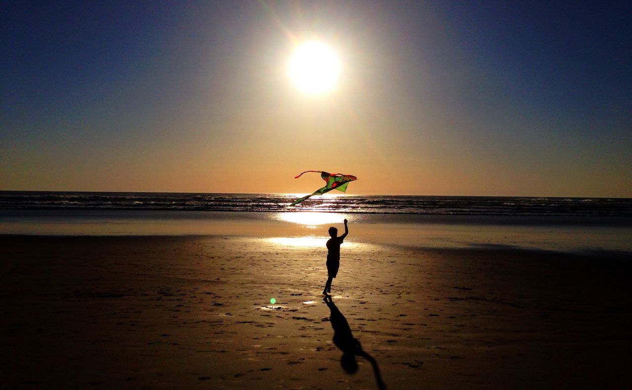 Silhouette boy flying kite at beach against sky during sunset