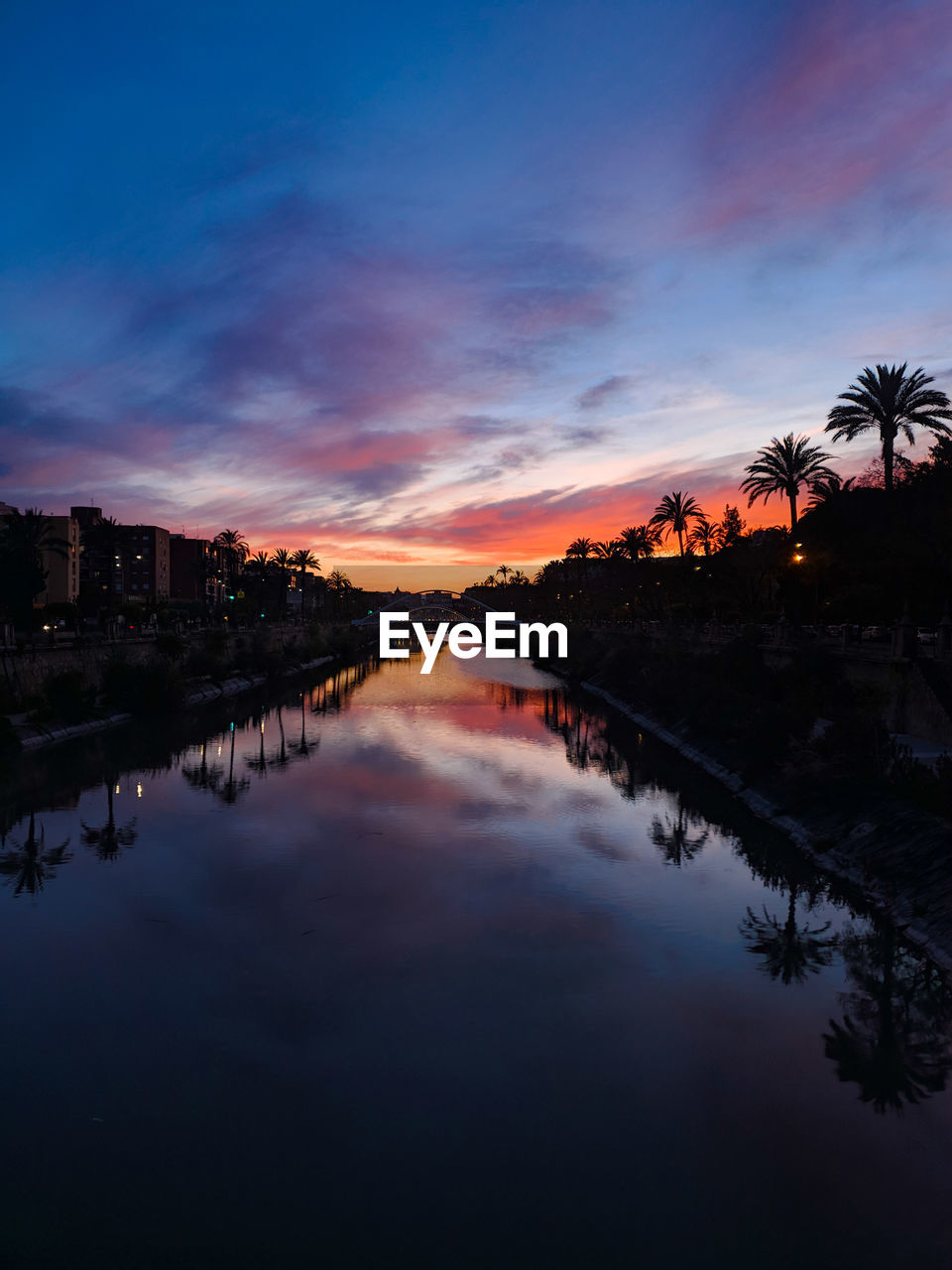 reflection, water, sky, dusk, evening, sunset, nature, cloud, tree, beauty in nature, architecture, scenics - nature, landscape, tranquility, horizon, environment, plant, travel destinations, no people, city, twilight, outdoors, tranquil scene, afterglow, river, blue, sunlight, tropical climate, built structure, travel, sun, land, palm tree, building, building exterior, silhouette, multi colored