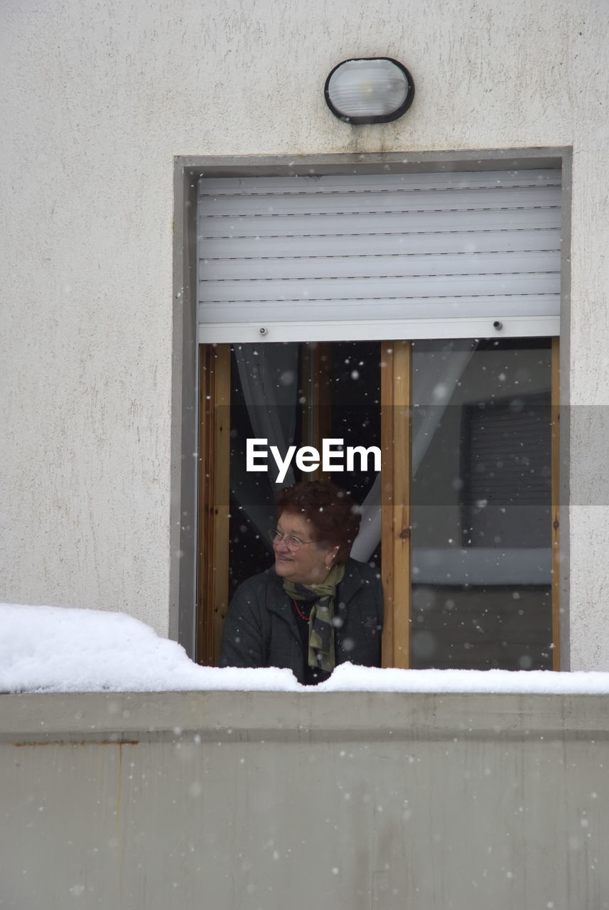 Woman looking away while standing by window during snowfall