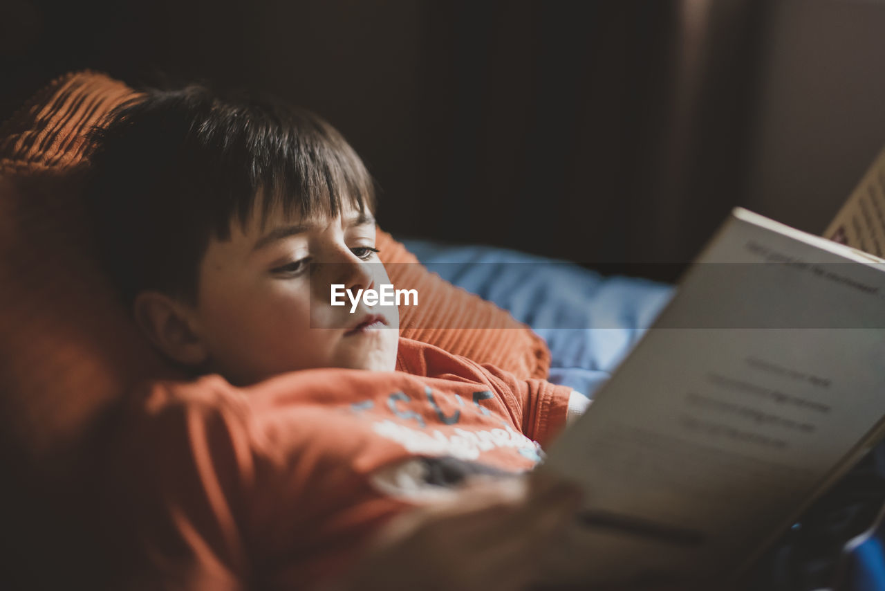 Boy reading book while lying on bed at home