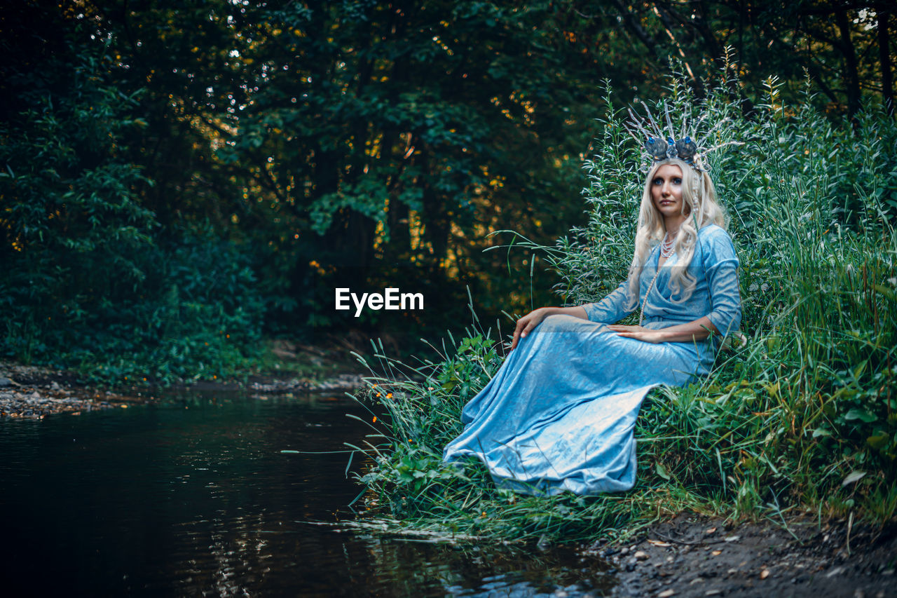 one person, women, adult, fashion, nature, plant, young adult, tree, dress, water, forest, hairstyle, blond hair, long hair, sunlight, full length, clothing, outdoors, fantasy, land, tranquility, green, beauty in nature, environment, darkness, portrait, female, sitting, elegance, fairy tale, lifestyles, mythical creature, contemplation, emotion, lake, day