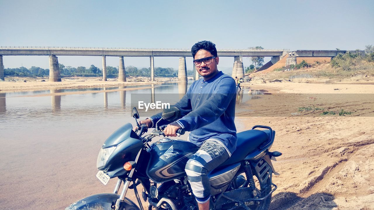 PORTRAIT OF SMILING MAN SITTING ON MOTORCYCLE AGAINST SKY