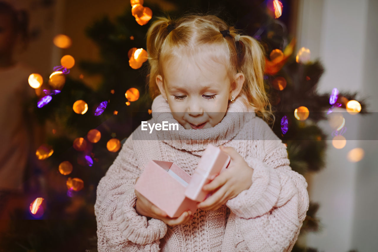 Cheerful baby girl with a gift box on the background of festive christmas tree on new year's eve