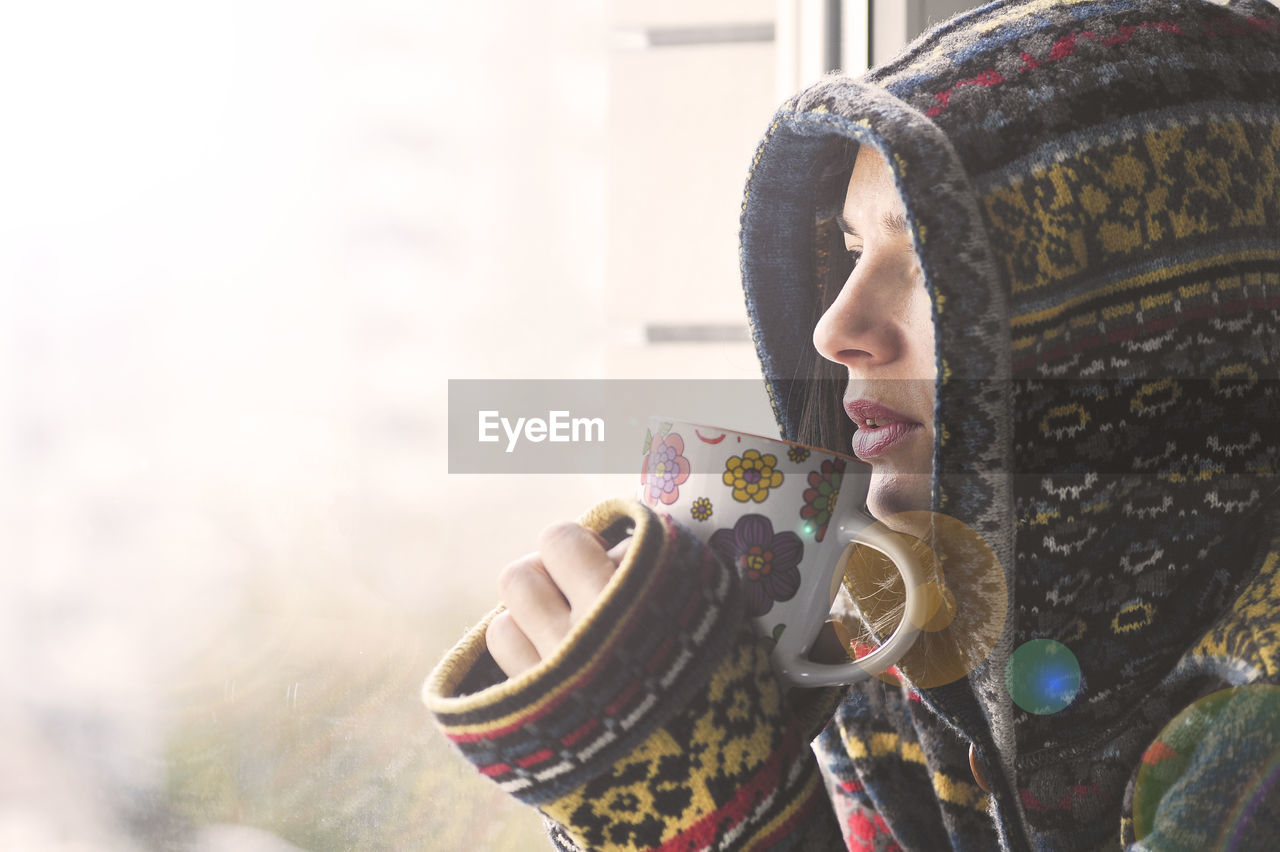 Woman wearing hooded jacket drinking coffee by window