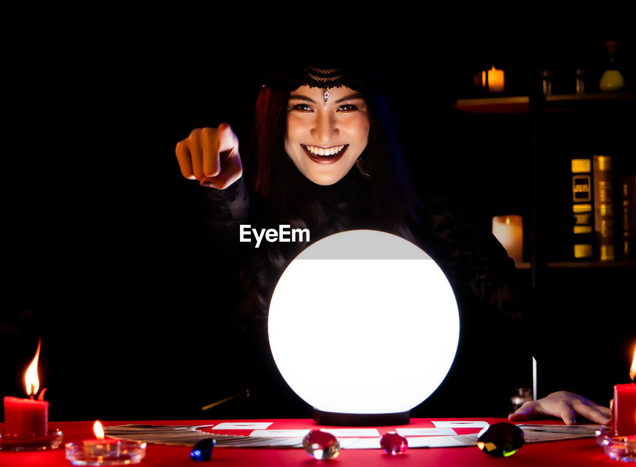Portrait of happy female fortune teller with illuminated crystal ball on table in darkroom