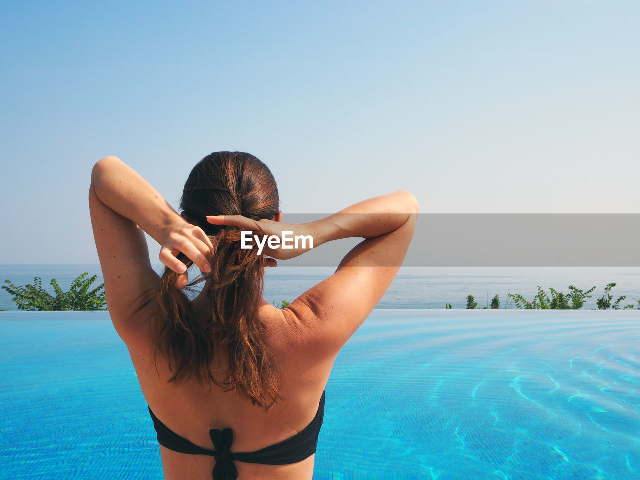 Rear view of woman adjusting hair by infinity pool against clear sky