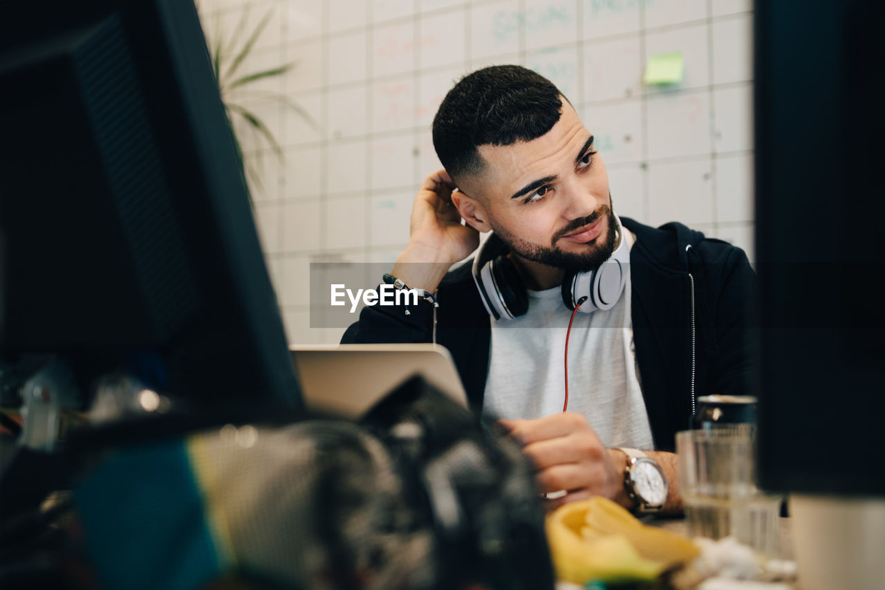 Confused young male computer programmer scratching head while sitting with laptop at creative office