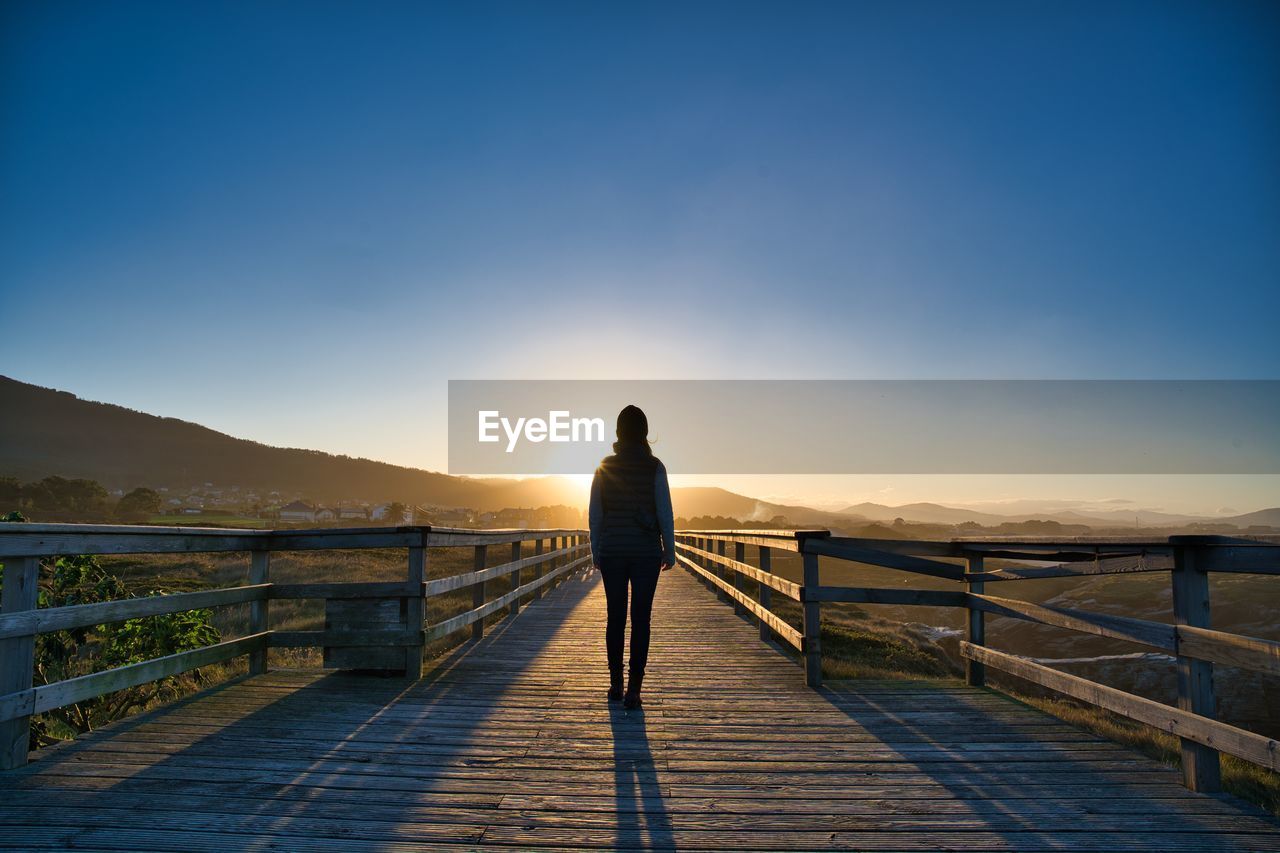 Rear view of woman standing on boardwalk against sky during sunset