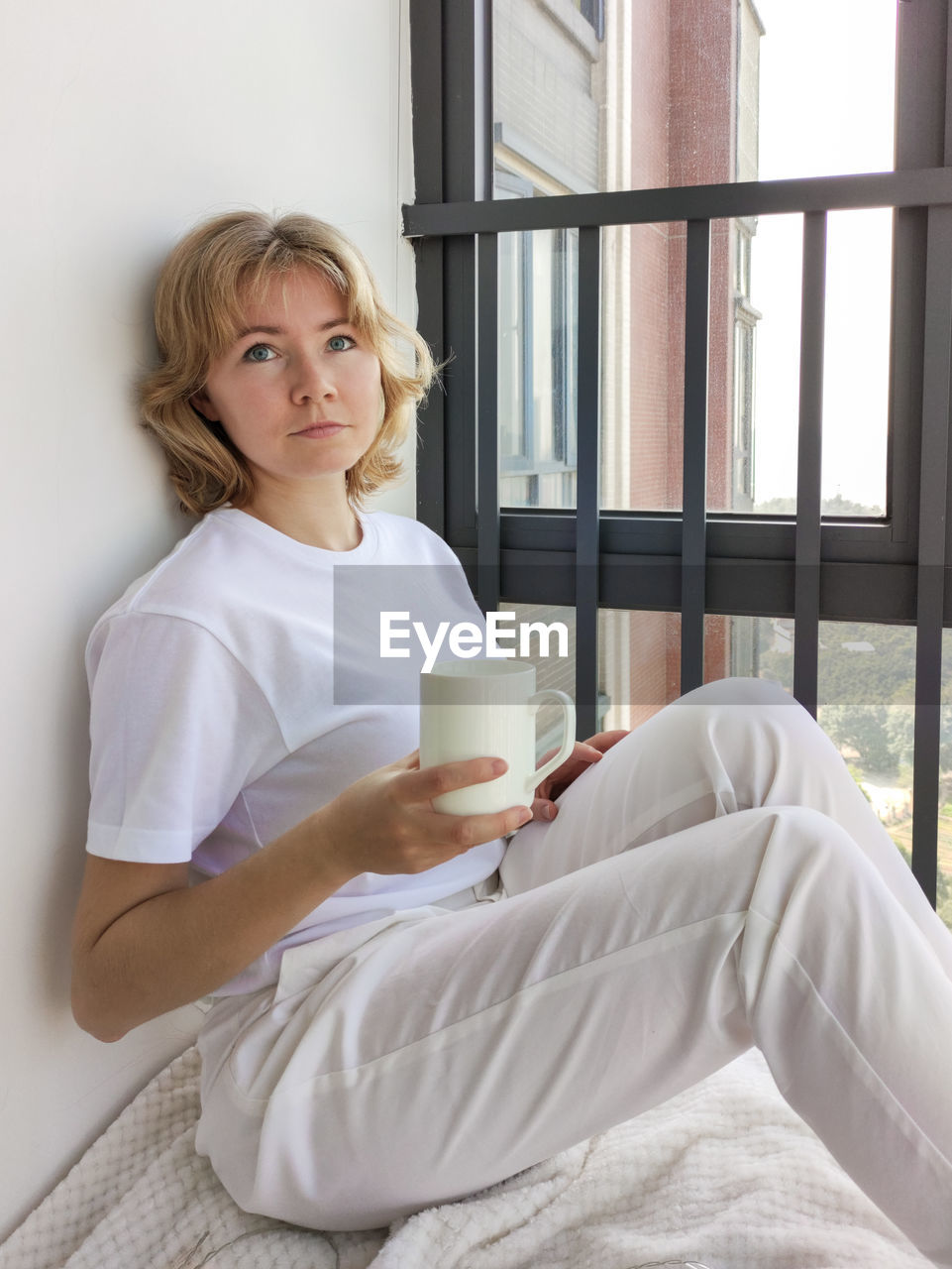 Attractive young sad woman sitting on the windowsill with a white cup in her hands