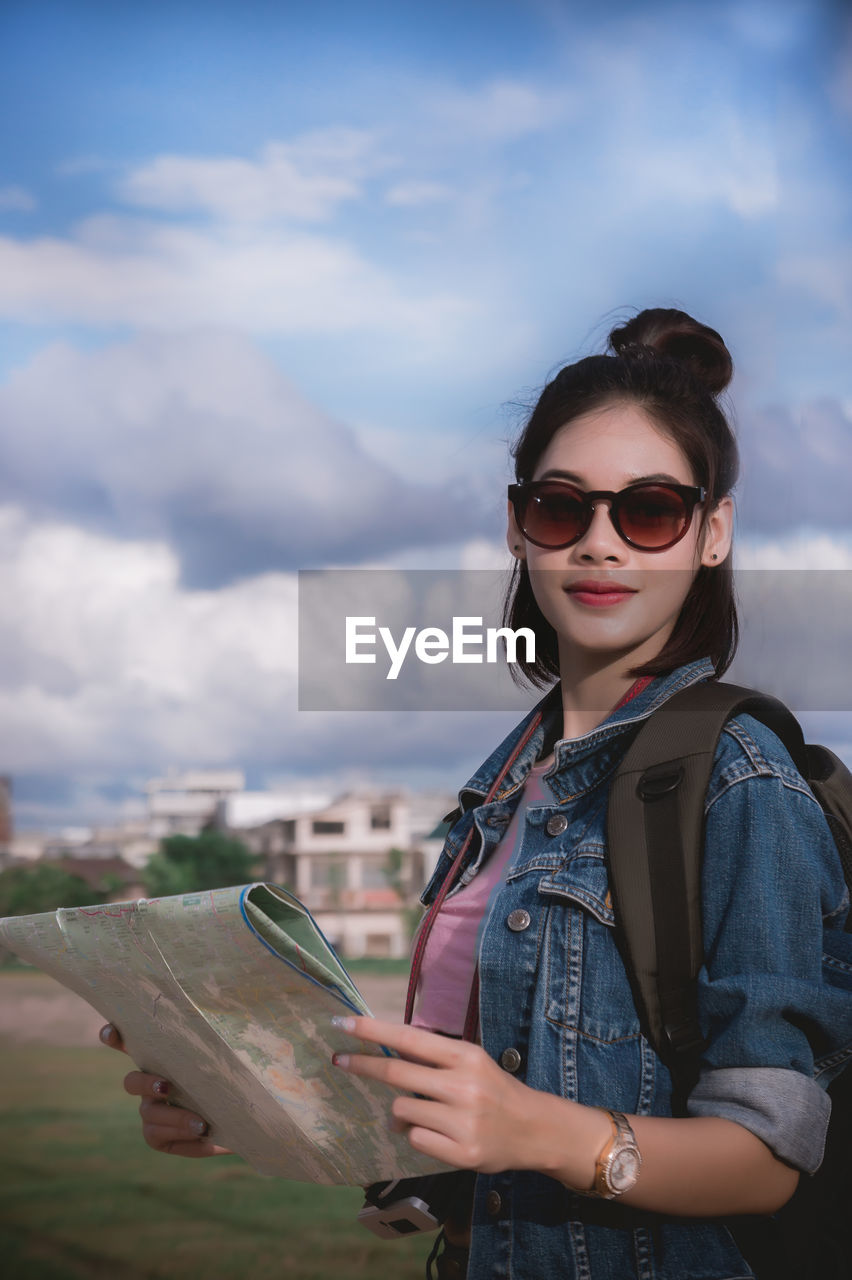 Portrait of young woman wearing sunglasses while holding map against sky