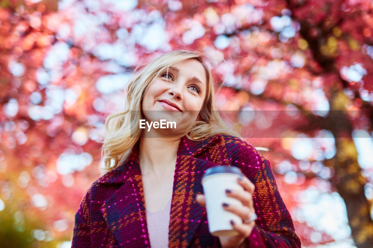 Young woman holding coffee while standing against autumn trees