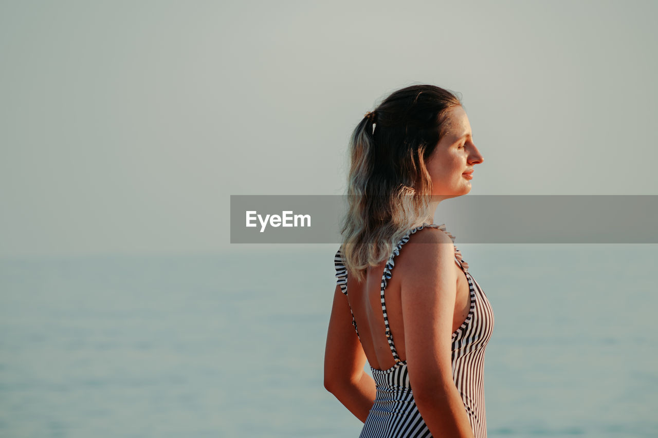 Side view of young woman standing in sea against sky