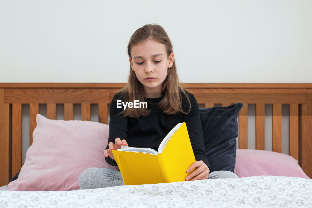 Young school age girl sitting on bed in bedroom and reading from yellow textbook. school homework.