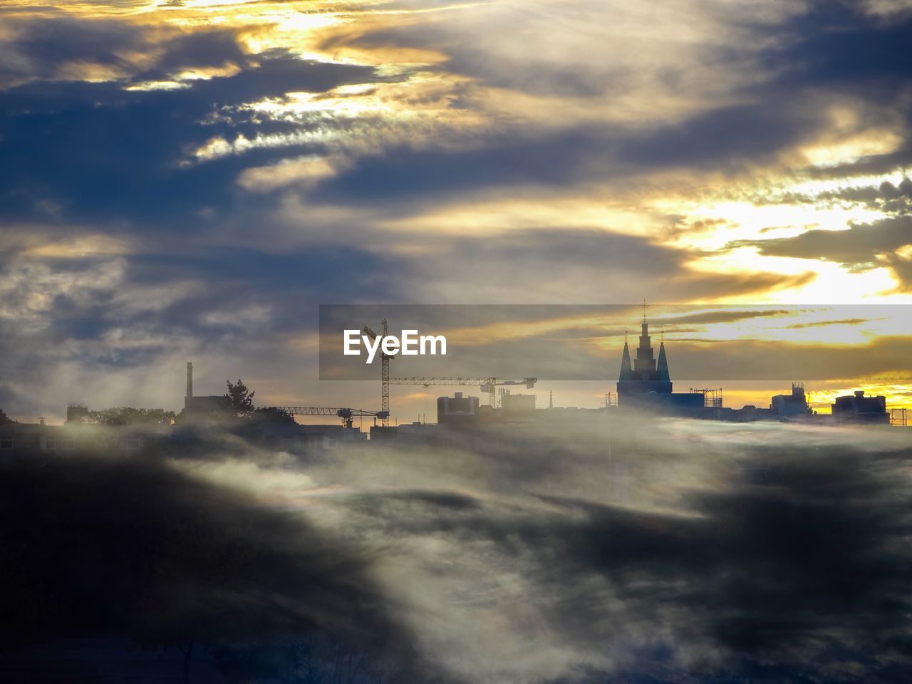 Buildings and cranes in foggy weather against cloudy sky during sunset