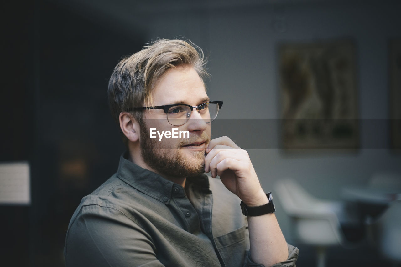 Thoughtful male computer programmer with hand on chin looking away in office