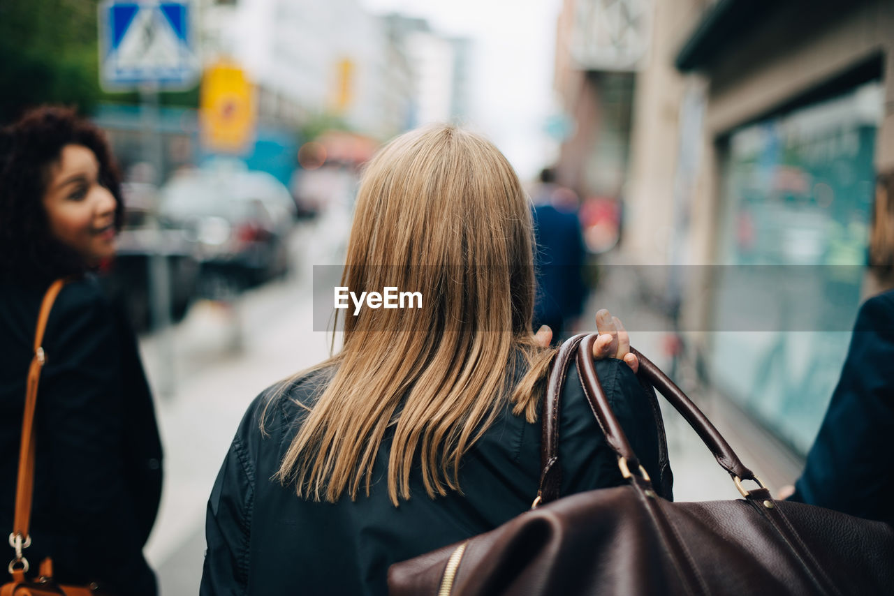 Rear view of businesswoman carrying luggage while walking amidst colleagues on sidewalk in city