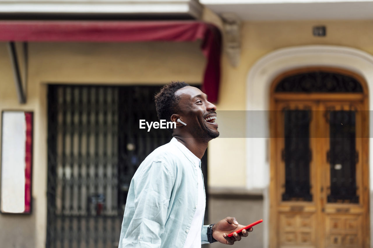 Happy young man with in-ear headphones holding smart phone at street