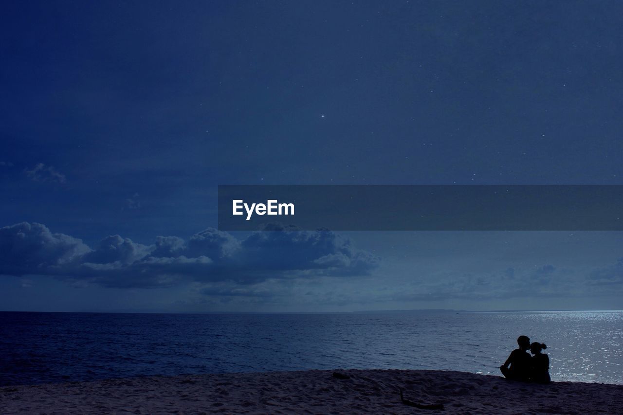 Silhouette couple sitting on beach against sky at night