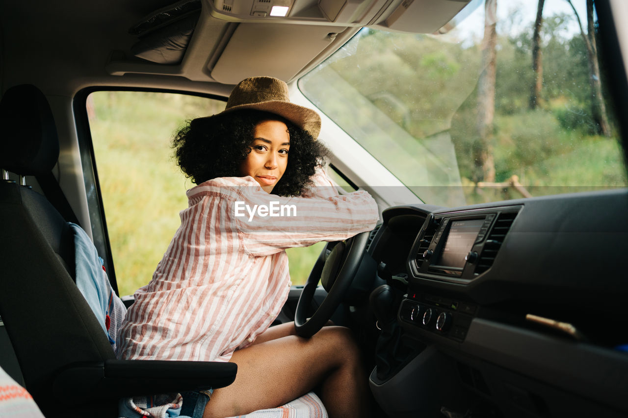Side view of romantic thoughtful young african american female traveler sitting on driver seat and looking at camera while enjoying summer journey in nature