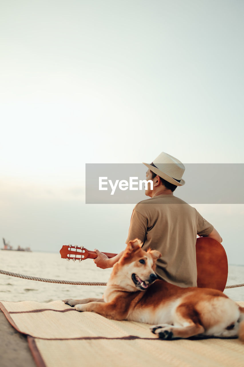 Rear view of man holding guitar sitting with dog against sky