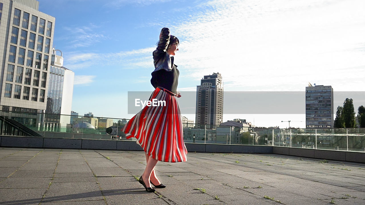 Happy woman wearing striped skirt and jacket on footpath in city