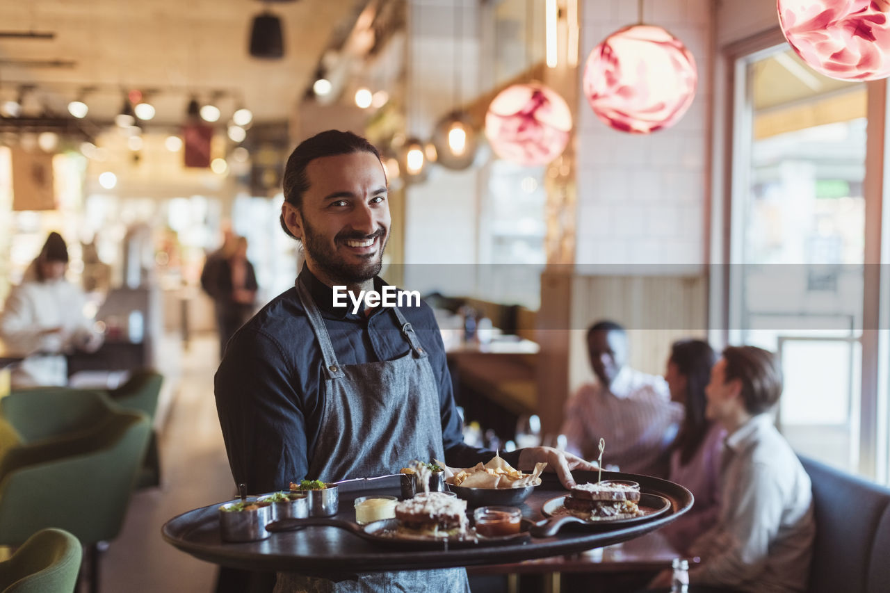 Portrait of smiling waiter with food while customer sitting in background at bar