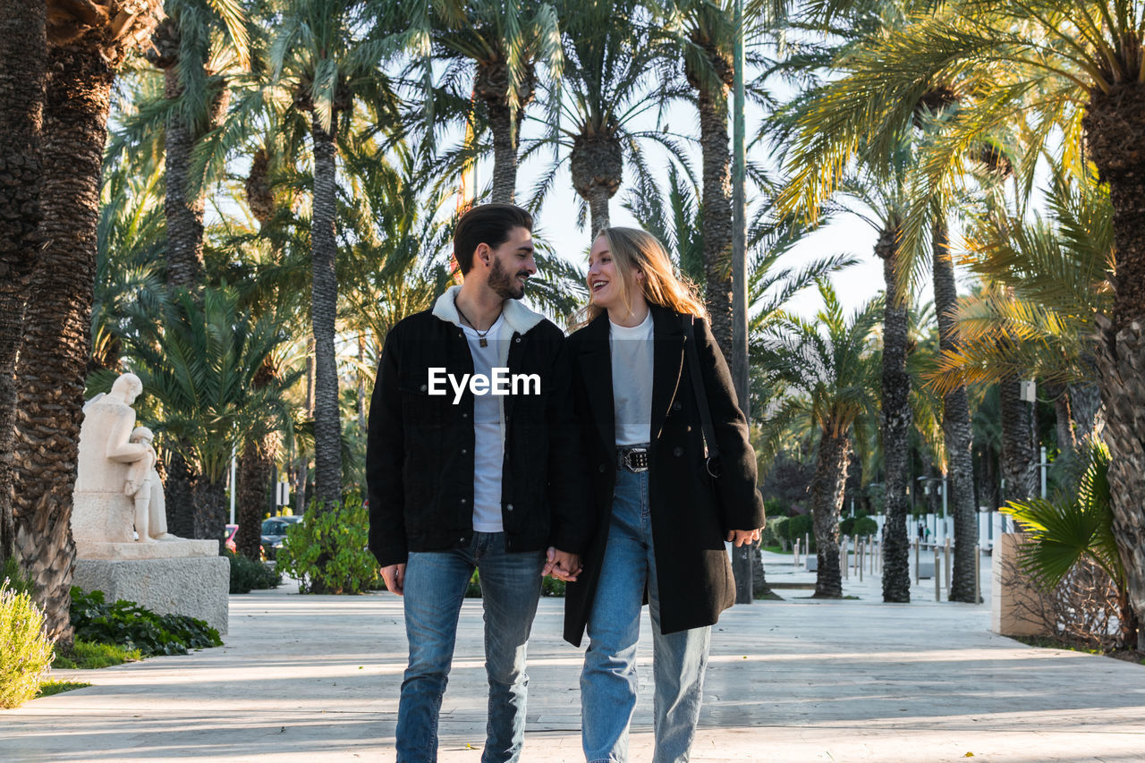 Cheerful young romantic couple in jeans and trendy black jackets walking on alley in park with tropical palms and enjoying holidays together in sunny day