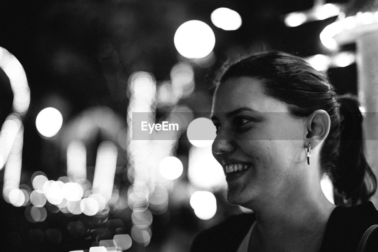 Close-up of smiling young woman looking away against illuminated lights