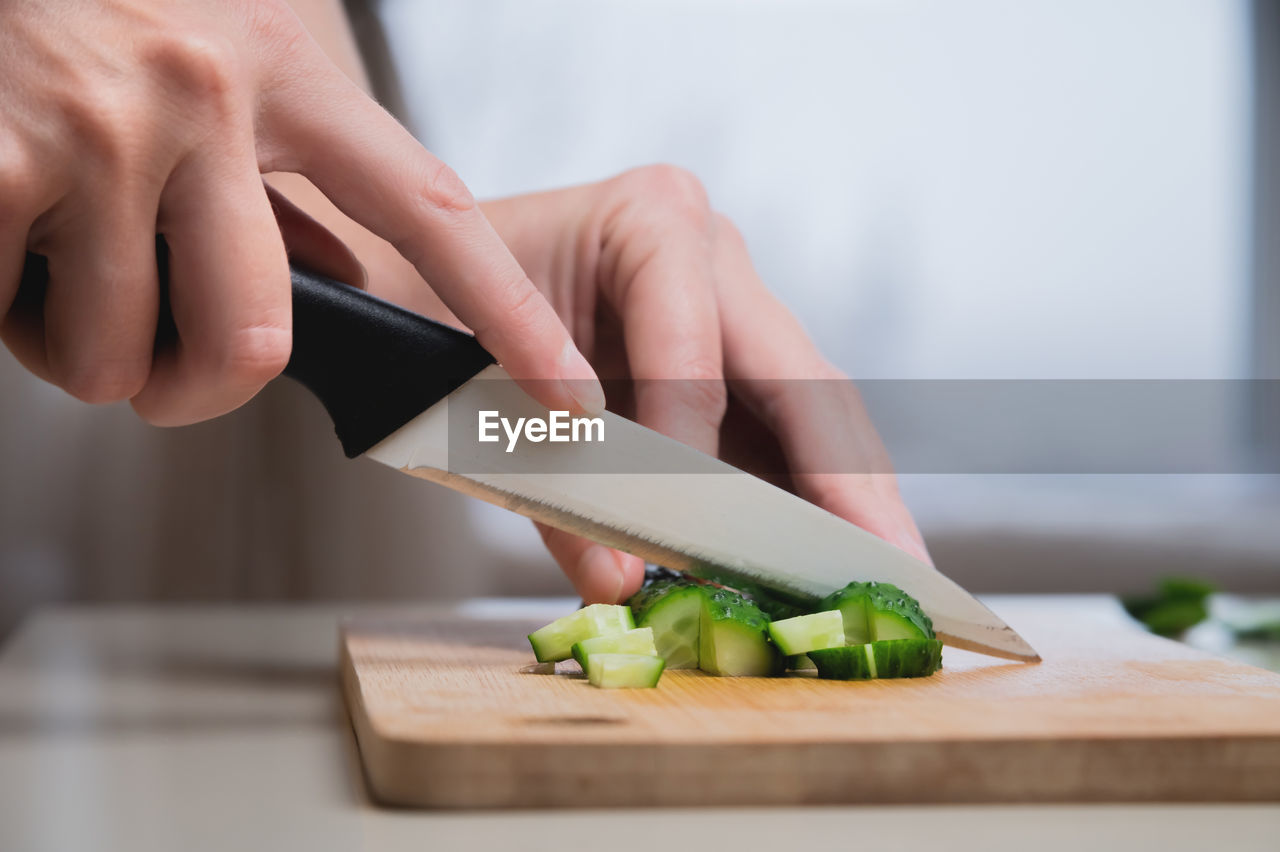 Close-up of female hands cutting on wooden cutting board fresh green cucumber for salad, home food