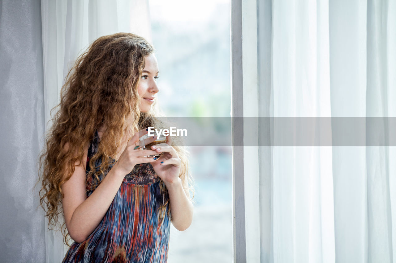 Woman having tea while looking through window while standing at home