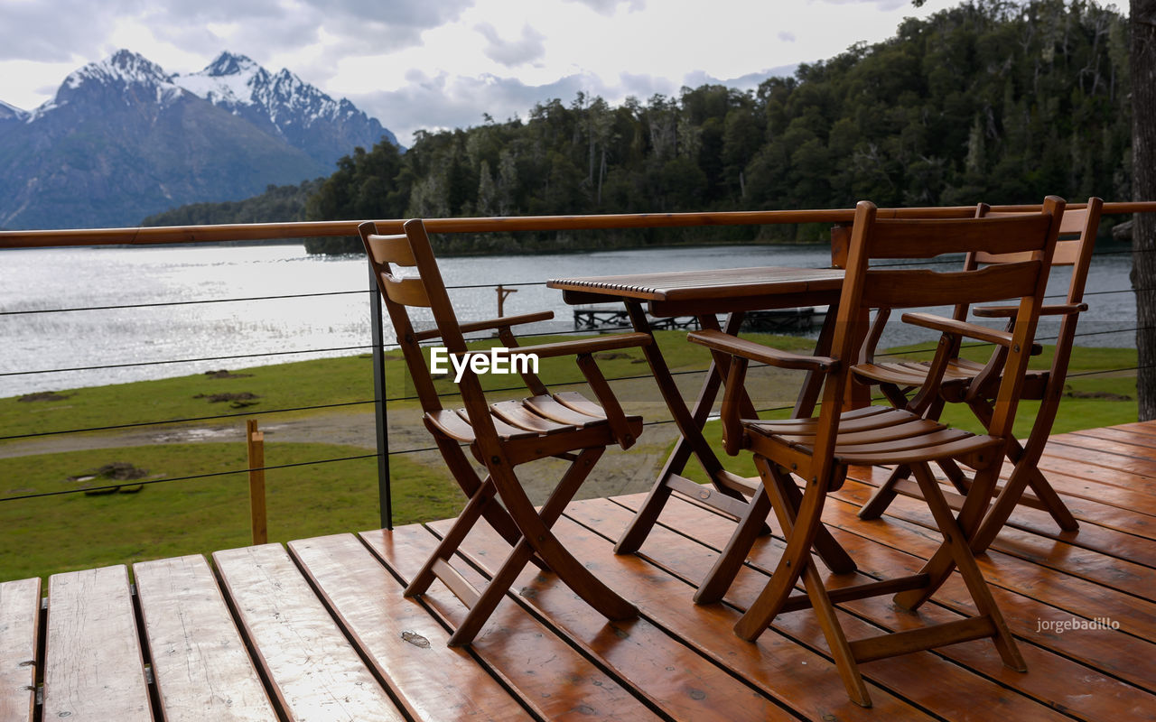 EMPTY CHAIRS AND TABLES BY LAKE AGAINST MOUNTAINS