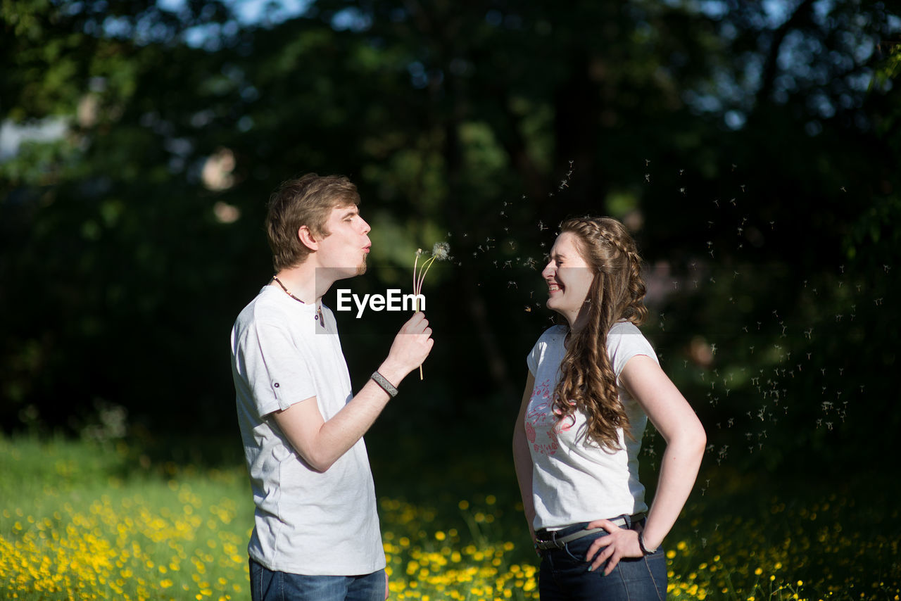 Man blowing dandelion seeds on cheerful girlfriend standing at park