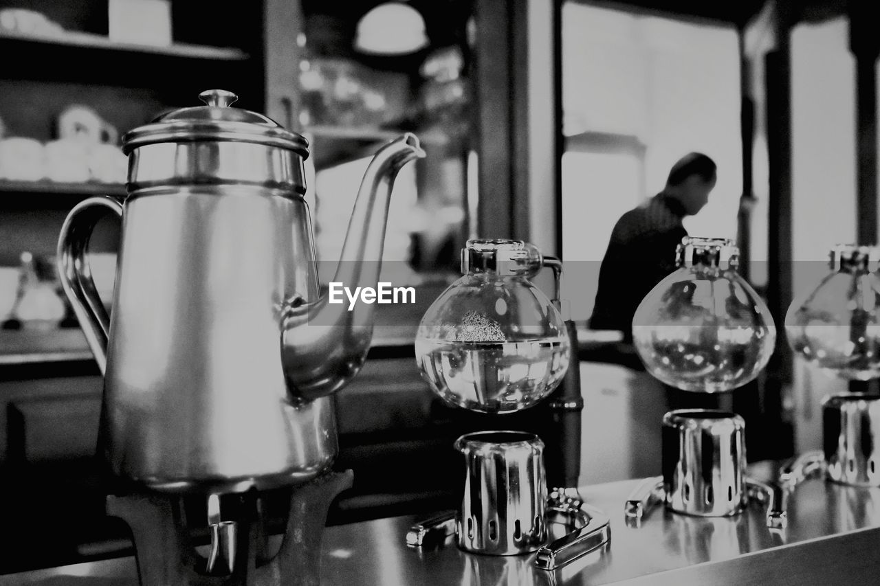 indoors, black, black and white, monochrome, food and drink, kettle, household equipment, monochrome photography, drink, domestic room, restaurant, kitchen utensil, kitchen, refreshment, person, table, focus on foreground, white, business, glass, cafe, coffeemaker