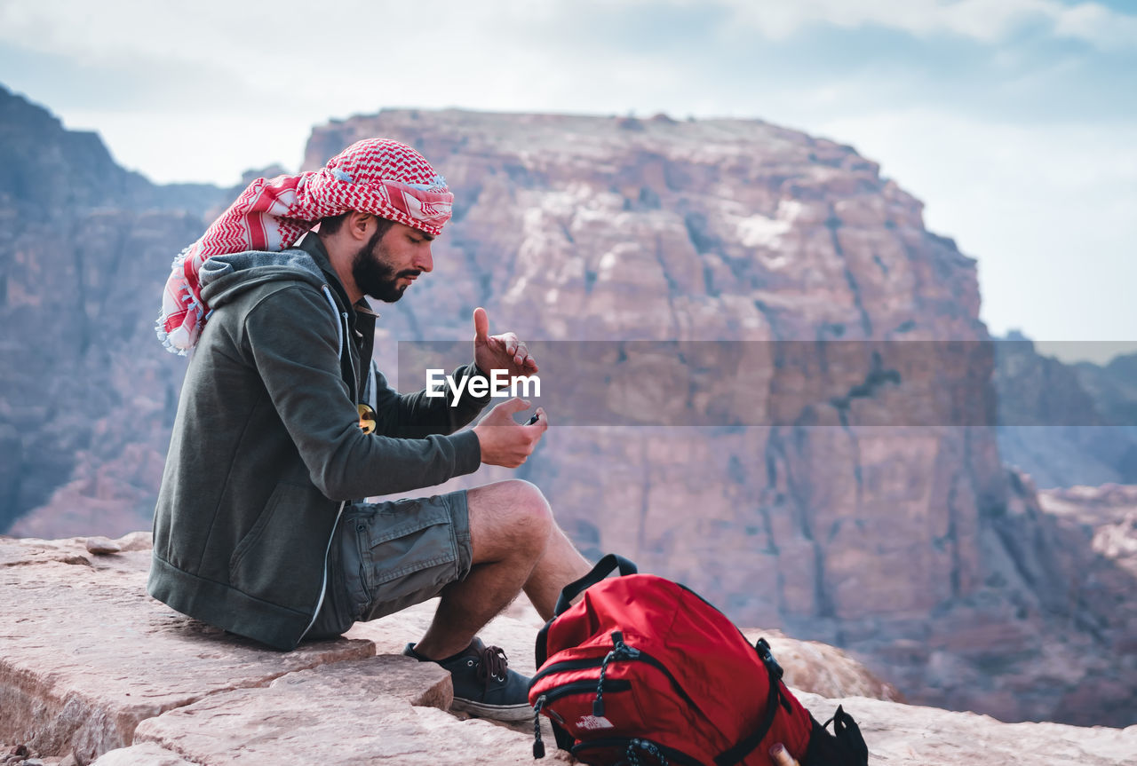 SIDE VIEW OF YOUNG MAN SITTING ON ROCK WITH MOUNTAIN