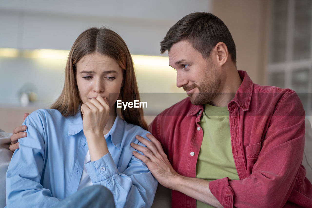 Crying, sad woman in depression while smiling husband lovingly hugs and calms her on sofa at home