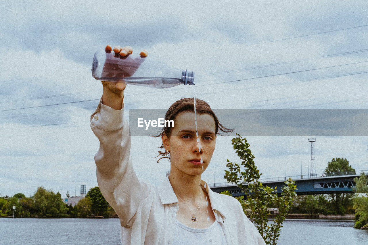 Portrait of confident young non-binary person pouring water from bottle against sky